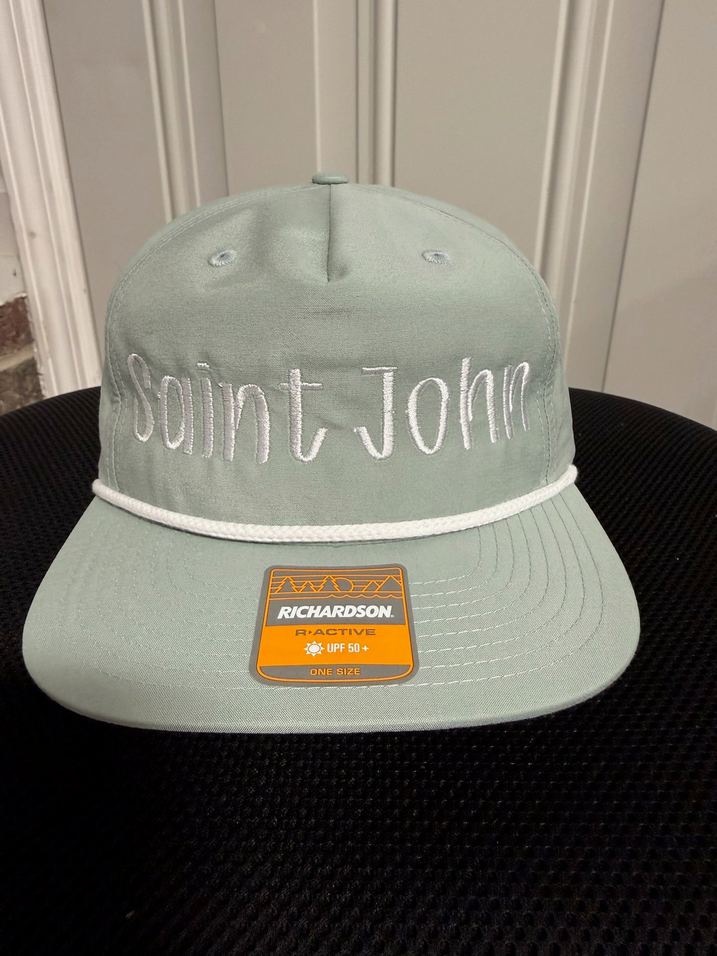 A light sage green flat-brim cap with 'Saint John' embroidered in white across the front, featuring a white rope detail and a prominent orange Richardson brand sticker on the brim.