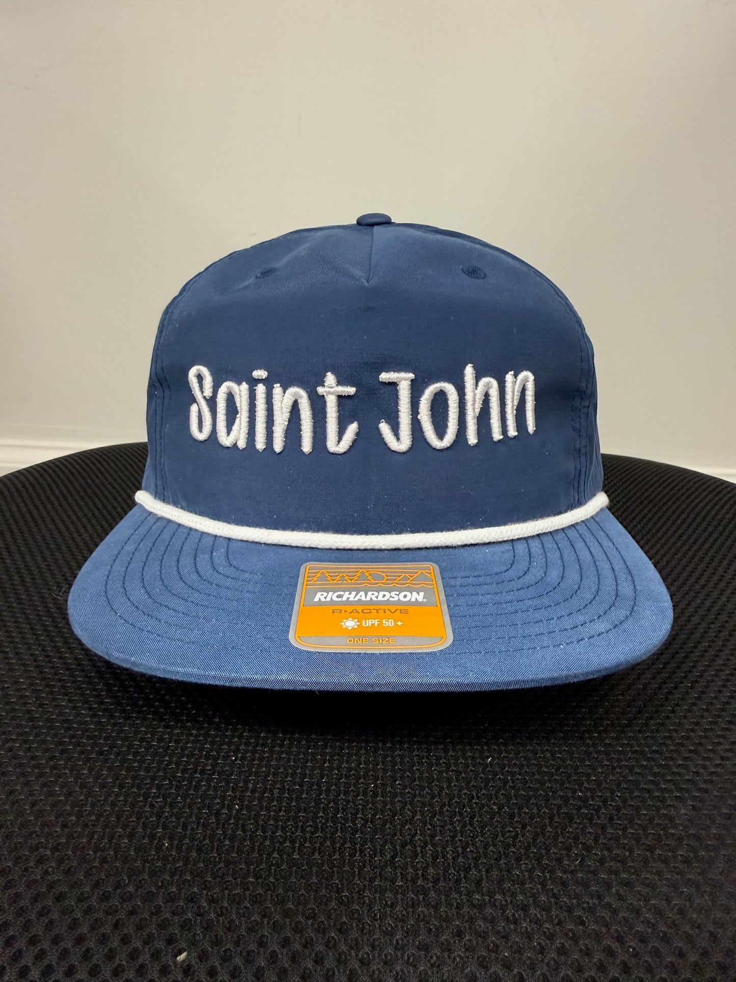 A blue flat-brim baseball cap with 'Saint John' embroidered in white across the front, a white stripe under the brim, and a visible Richardson brand patch on the brim.