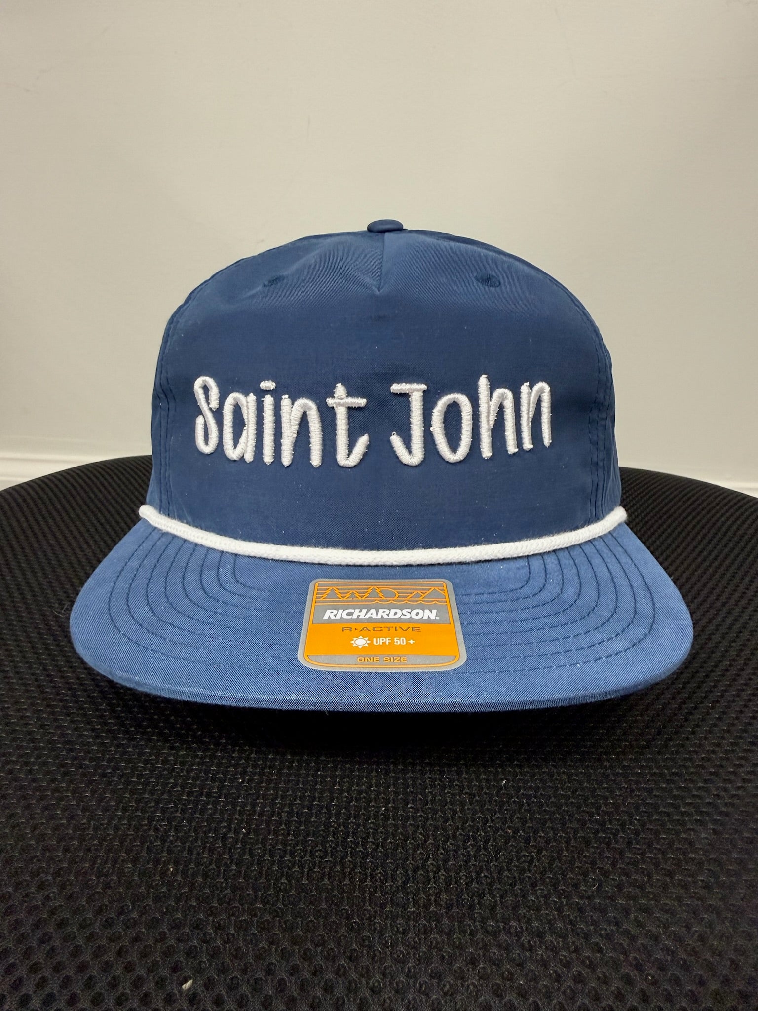 A blue flat-brim baseball cap with 'Saint John' embroidered in white across the front, a white stripe under the brim, and a visible Richardson brand patch on the brim.